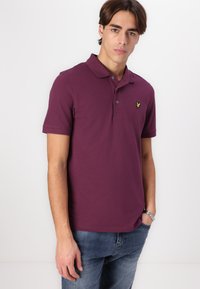 Polo tričko v tmavosfioletovej bavlne s krátkymi rukávmi, rebrovaným golierom a gombíkovou légou. Na hrudi má malé žlté logo.