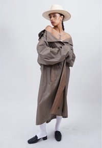 Frau in einem übergroßen braunen Trenchcoat, der über die Schultern fällt, weißen Socken, schwarzen Schuhen und einem Strohhut, stehend vor einem schlichten hellen Hintergrund.