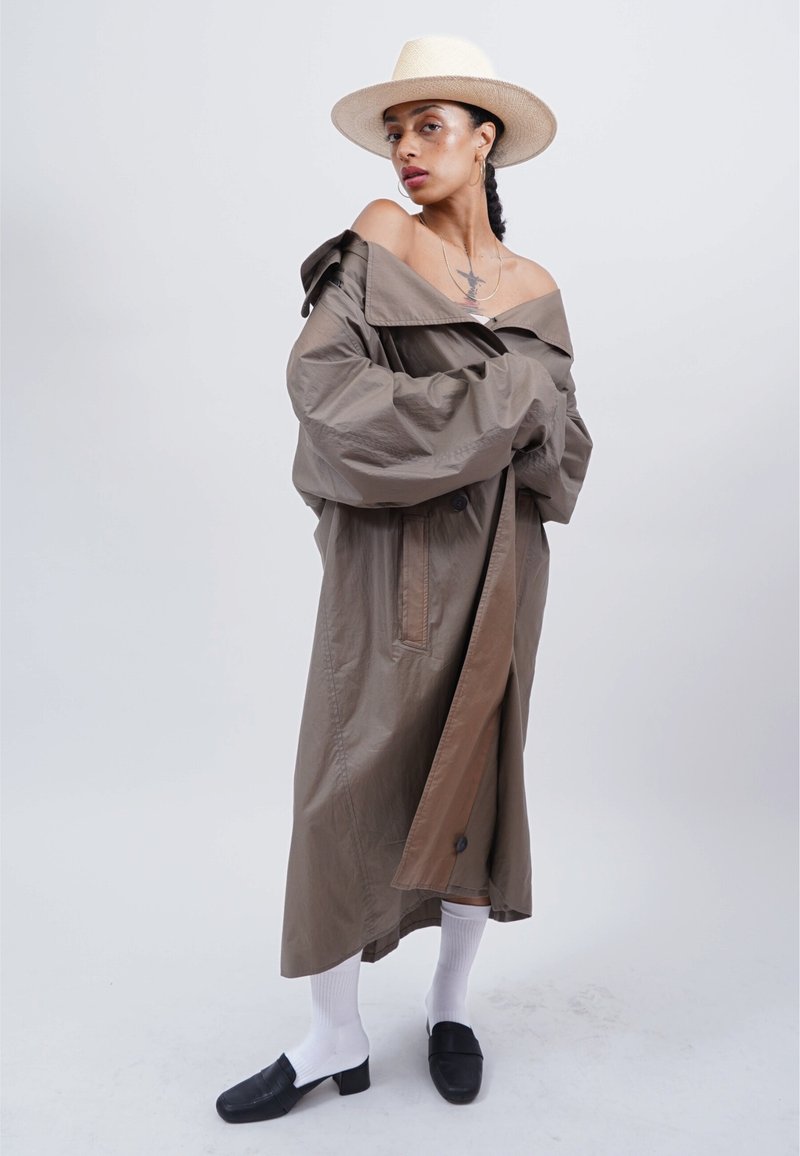 Frau in einem übergroßen braunen Trenchcoat, der über die Schultern fällt, weißen Socken, schwarzen Schuhen und einem Strohhut, stehend vor einem schlichten hellen Hintergrund.