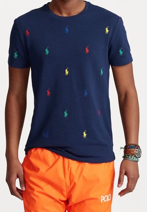 Homme portant un T-shirt bleu marine avec des logos multicolores de joueurs de polo et un short orange vif avec un logo blanc "POLO".