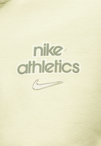 Κείμενο Nike Athletics και ασημί λογότυπο Nike swoosh σε φόντο από ανοιχτό μπεζ ύφασμα.