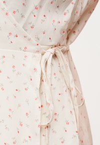 Κοντινή λήψη κρεμ υφασμάτινου φόρεματος τύπου wrap με μικρό σχέδιο από κόκκινες και ροζ τριανταφυλλιές, δεμένο στη μέση με φιόγκο από ύφασμα.