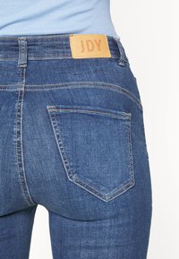 Jeans de ganga em azul escuro com uma textura suave. As características incluem dois bolsos traseiros e um rótulo de couro bege com a gravação "JDY".