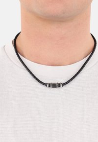 Collana intrecciata in pelle nera con un design centrale di perline e accenti argentati, con piccoli dettagli neri, indossata su una camicia bianca con texture.
