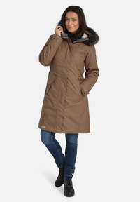 Bruine geïsoleerde parka met een capuchon met faux fur, ritssluiting en zijzakken. Draagt over een zwarte top en een blauwe spijkerbroek, met zwarte laarzen.