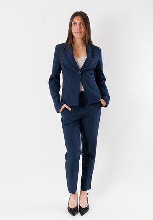 Giacca di abito blu navy su misura con chiusura a un bottone, accompagnata da pantaloni tapered coordinati. Tessuto liscio, design elegante, accenti minimali.