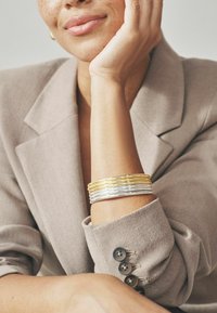 Metallist käevõru, millel on vahelduvad kuld- ja hõbetoonid, asetatud pehme, heleda beežiga bleiseri taustale, millel on nööbid.