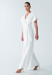 Witte omslagjurk met korte, wapperende mouwen, gestrikt in de taille. Gladde stof, ontwerp tot op de vloer. Gecombineerd met gouden sandalen met hak en bandjes.