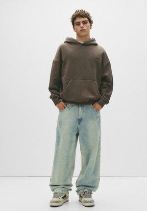 Brown hoodie with a front pocket, paired with light blue, oversized jeans. The outfit is completed with grey and white sneakers.