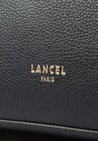 Fekete textúrájú bőr aranybélyegzett "LANCEL" és "PARIS" márkajelzéssel. Diszkrét szemcsés minta jellemzi, amely fokozza a felszíni részleteket.