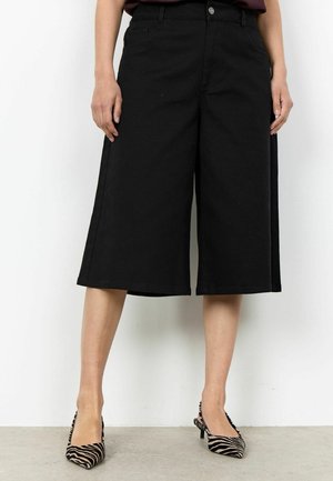 Femme portant un short noir large mi-cuisse et des talons pointus imprimé zèbre, debout contre un mur blanc.