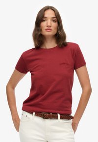Rode katoenen t-shirt met korte mouwen en ronde hals, met een subtiele geborduurde logo op de linkerborst. Gecombineerd met witte jeans en een bruine gevlochten riem.