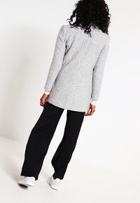 Abrigo de mezcla de lana gris con superficie texturizada, mangas largas y diseño recto, combinado con pantalones de pierna ancha negros y deportivas blancas.