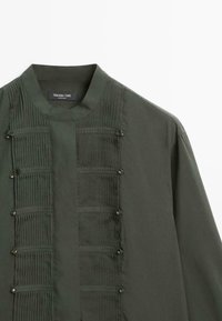 Chemise à manches longues vert foncé avec col montant et panneaux plissés verticaux à l'avant, agrémentée de petits clous métalliques de chaque côté.