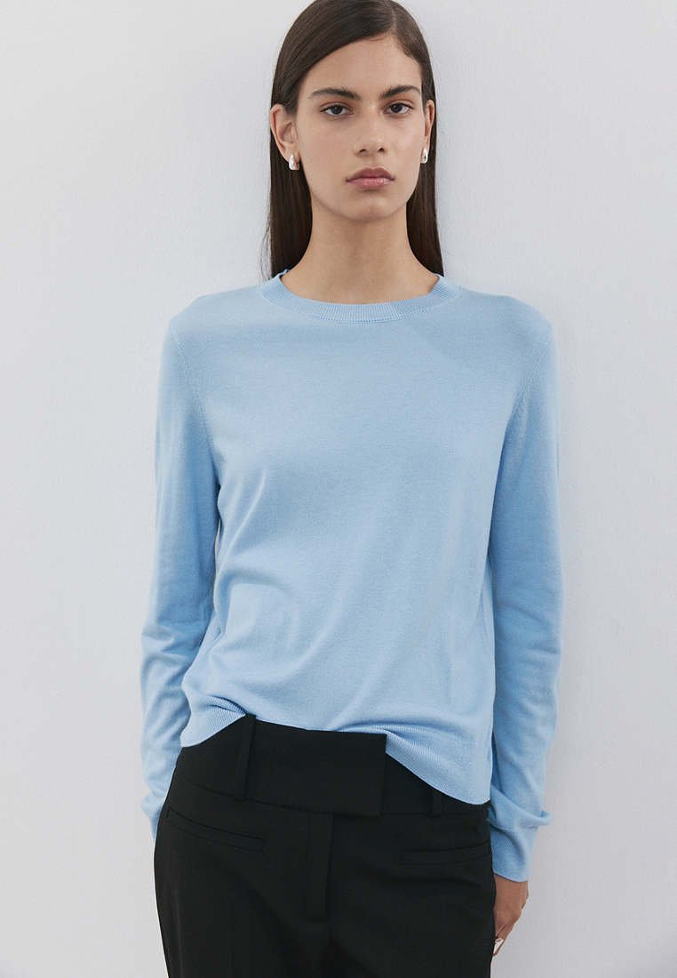 Jeune femme aux longs cheveux foncés portant un pull bleu pâle et un pantalon noir, debout devant un fond clair uni avec une expression neutre.