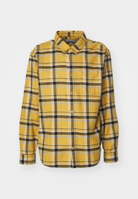 Camisa a cuadros amarilla con acentos en negro y azul, de mangas largas, cuello abotonado y un bolsillo en el pecho. Textura de tela suave.