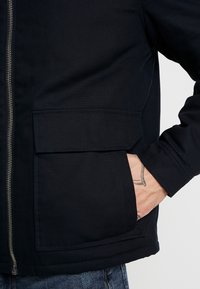 Mörkblå, zippered jacka gjord av texturerad tyg. Har en bröstficka och sidfickor. Manschetterna är avslutade med en sturdy fåll.