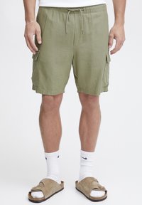 Shorts cargo vert olive avec deux poches latérales, taille élastique et cordon de serrage. Le tissu a une surface texturée et une coupe décontractée.