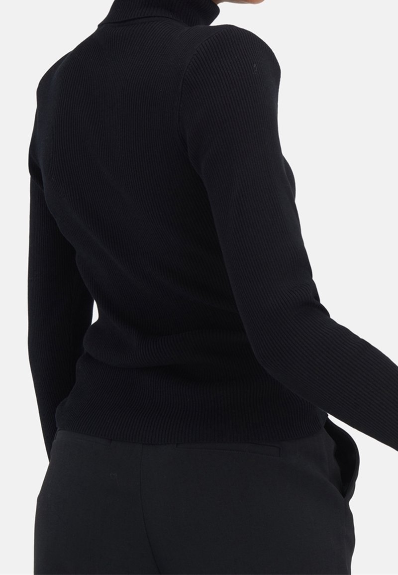 Personne portant un pull à col roulé côtelé noir ajusté et un pantalon noir, vue de dos et de côté, une main dans la poche.