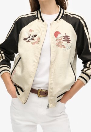 Femme portant une veste bomber en satin crème et noire avec des motifs brodés de grue, fleurs de cerisier, montagne, soleil et pagode.