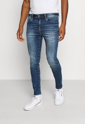 Mężczyzna ubrany w dopasowane niebieskie dżinsy i białe buty sportowe Adidas, stojący na tle jednolitego jasnego tła.