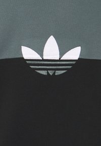 Černá a šedá bavlněná tkanina s výrazným logem Adidas, které zobrazuje tři listy a tři vodorovné pruhy, vyšitá bílou barvou.