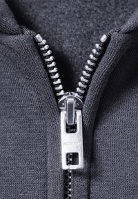 Lue med glidelås laget av mørkegrå stoff, glatt tekstur og en metallisk sølvfarget glidelås med en merket trekant, delvis åpnet.