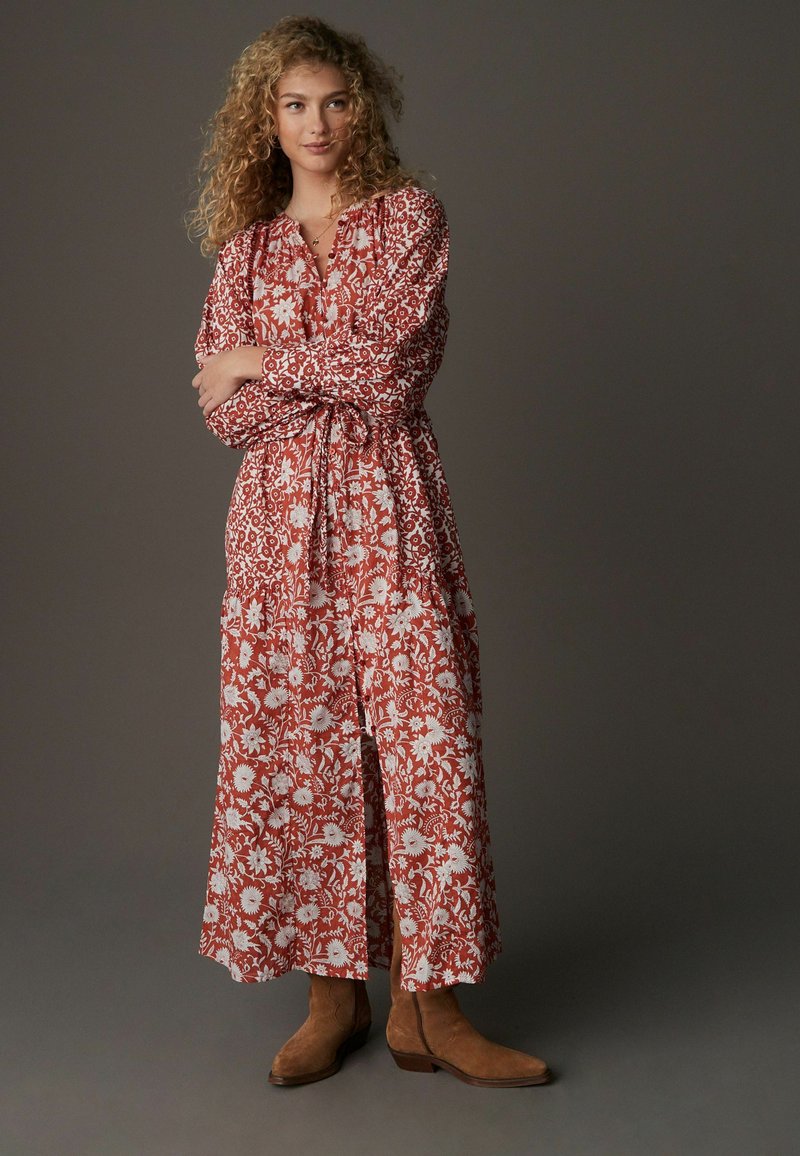Rochie maxi roșie cu flori și modele albe, cu mâneci lungi, guler adunat și curea. Asortată cu bocanci bej până la gleznă. Textură de stofă netedă.