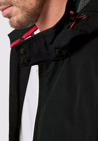 Veste noire avec un col haut et un accent rouge, boutons-pression et intérieur texturé. Détail de couture visible le long des bords.