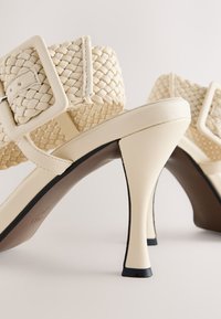 Sandales à talons hauts beiges avec un matériau tressé sur les brides, une finition en cuir lisse, et un talon fin avec un accent noir à la base.