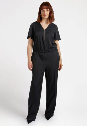 Zwarte jumpsuit met korte mouwen, V-hals, ritssluiting aan de voorkant, elastische tailleband en brede, vloeiende broekspijpen. Textuur zwarte hakken maken de look compleet.