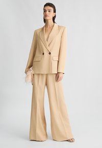 Blazer beige su misura con chiusura doppiopetto e rever a punta, abbinato a pantaloni a gamba larga. Include una borsa a tracolla decorativa con piume.