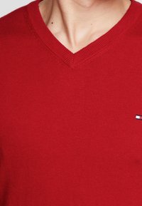 Mies, joka pukeutuu punaiseen V-pääntie-neuleeseen, jossa on pieni suorakulmainen logo oikeassa rintataskussa.