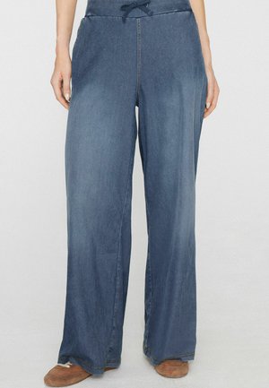 Frau trägt weite, locker sitzende blaue Jeans mit Kordelzug an der Taille, steht mit entspannt an den Seiten hängenden Händen, braune Schuhe sichtbar.