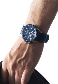 Mėlynas analoginis laikrodis su sidabro akcentais, dideliais skaitmenimis, datos langeliu ir tekstūruotu juodu guminiu dirželiu ant riešo.