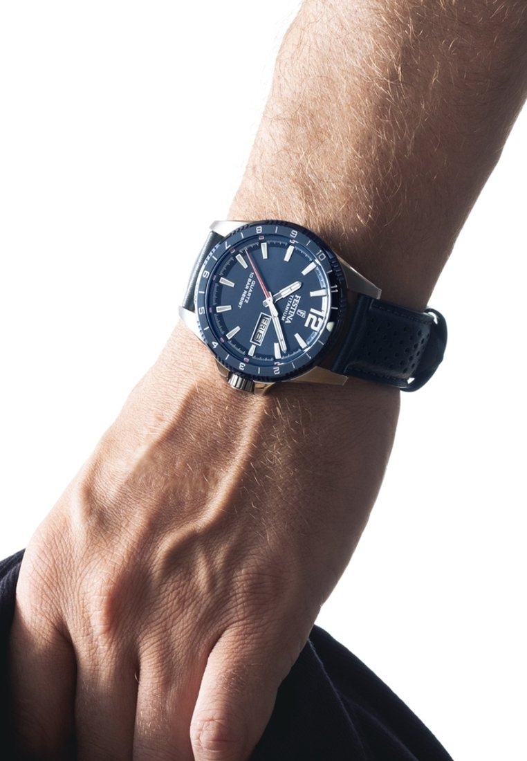 Mėlynas analoginis laikrodis su sidabro akcentais, dideliais skaitmenimis, datos langeliu ir tekstūruotu juodu guminiu dirželiu ant riešo.