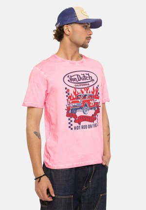 Homme portant un T-shirt rose Von Dutch avec une voiture rouge vintage, des flammes et le texte « Hot Rod on Fire », associé à un jean foncé et une casquette bleue.
