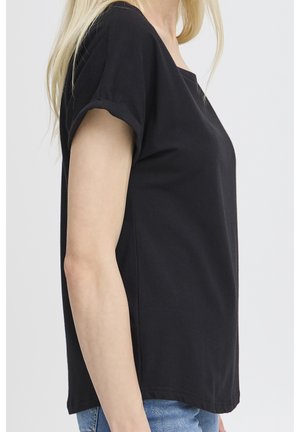 Lyshåret kvinde iført en ensfarvet sort T-shirt med korte ærmer og blå jeans, set fra siden mod en ensartet baggrund.