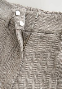 Primer plano de la pretina de tejido en espiga beige y blanco con cremallera y dos cierres de gancho y ojal plateados en pantalones o falda.