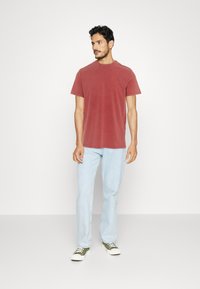 Maglietta rossa a maniche corte con collo rotondo, abbinata a jeans blu chiaro e sneakers verdi. Dettagli di cucitura minimi sulla maglietta.