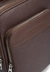 Καφέ δερμάτινη τσάντα με υφασμάτινο σχέδιο, διακοσμημένη με ασημένια φερμουάρ και το ανάγλυφο λογότυπο "TOMMY HILFIGER" στο μπροστινό μέρος. Συμπαγής και ανθεκτική.