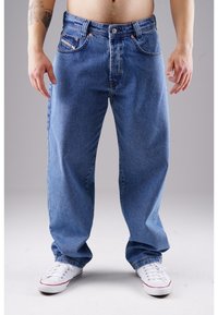 Hellblaue Jeans mit entspannter Passform, fünf Taschen-Design, orangefarbene Nähte und Metallbeschläge. Getragen mit weißen Turnschuhen.