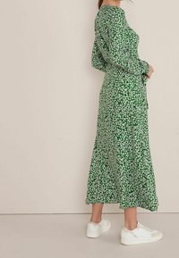 Robe florale verte avec des manches longues, une jupe fluide et des baskets blanches. Le tissu présente de petites fleurs blanches sur un fond vert vibrant.