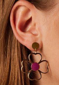 Gouden oorbellen in de vorm van een bloem met een paarse kern, met een cirkelvormig studs ontwerp. De oorbellen hangen onder de oorlel tegen bruin haar.