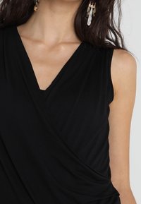 Top senza maniche nero con design avvolto, scollatura a V e tessuto morbido con drappeggi sottili. Accentuato da orecchini di colore chiaro.