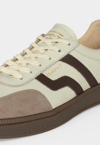 Baskets crème et marron avec bout en daim beige, semelle marron texturée, lacets crème et logo « GANT » sur le côté et la languette en lettres dorées.