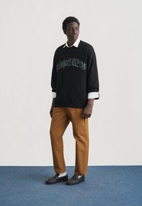 Άνδρας που στέκεται σε μπλε χαλί, φοράει μαύρο πουλόβερ πάνω από λευκό πουκάμισο, καφέ παντελόνι και μαύρα παπούτσια χωρίς κορδόνια, κοιτάζει προς τα πλάγια.