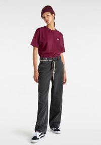 Personne portant un bonnet bordeaux, un t-shirt bordeaux, un pantalon noir à jambes larges avec une ceinture cordon blanche et des baskets noires et blanches, debout devant un fond blanc.