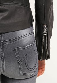 Gros plan d'une personne portant un jean en denim gris avec une poche arrière cousue et une veste en cuir noire avec une manche zippée.