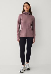 Giacca sportiva viola con colletto alto, zip frontale e silhouette aderente, abbinata a leggings neri e sneakers grigie.